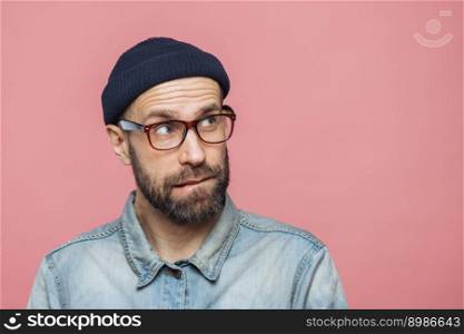 Portrait of unshaven thoughtful middle aged male with pensive expression looks upwards, wears stylish hat, eyewear and denim jacket, isolated over pink background with copy space for your text
