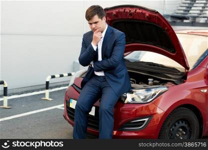 Portrait of upset businessman sitting on bonnet of broken car