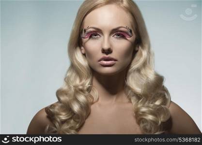 portrait of very pretty blonde girl with creative feathered make-up, shiny decorations on the face and long silky wavy hair-style. Looking in camera