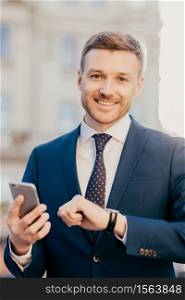 Portrait of well dressed handsome cheerful male executive manager checks time on his wristwatch, uses smart phone, checks email box and waits for bank confirmation, poses outdoor. Vertical shot
