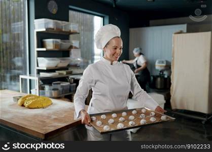Portrait of woman baker in uniform carrying tray with cookies shaped from dough and prepared for baking. Pastry production. Woman baker carrying tray with cookies shaped from dough
