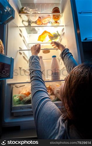 Portrait of woman in pajamas reaching donut on top shelf of fridge at night
