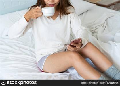 Portrait of woman using mobile phone and drinking coffee while sitting on the bed in the morning,close up