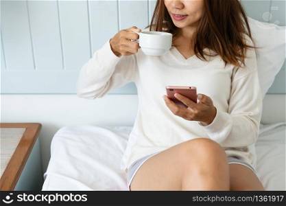 Portrait of woman using mobile phone and drinking coffee while sitting on the bed in the morning,close up