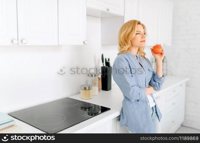 Portrait of woman with apple poses on the kitchen with snow-white interior. Female person at home in the morning, healthy nutrition and lifestyle. Woman poses on kitchen with snow-white interior