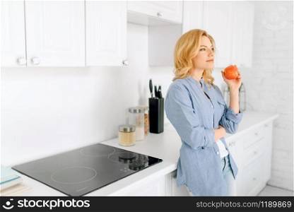 Portrait of woman with apple poses on the kitchen with snow-white interior. Female person at home in the morning, healthy nutrition and lifestyle. Woman poses on kitchen with snow-white interior