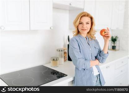 Portrait of woman with apple poses on the kitchen with snow-white interior. Female person at home in the morning, healthy nutrition and lifestyle