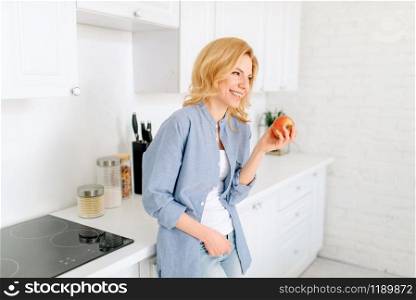 Portrait of woman with apple poses on the kitchen with snow-white interior. Female person at home in the morning, healthy nutrition and lifestyle. Woman poses on kitchen with snow-white interior