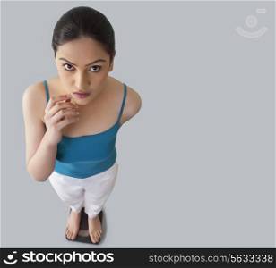 Portrait of worried young woman standing on weighing scale against gray background