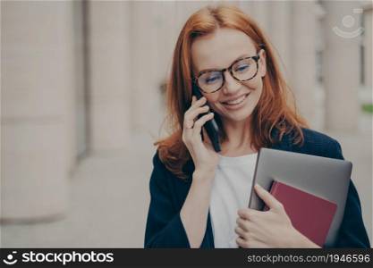 Portrait of young 30s caucasian business woman standing outdoors and talking on smartphone with client, arranging meeting with partner on mobile phone with smile on face, blurred city background. Young 30s caucasian business woman standing outdoors and talking on smartphone with client