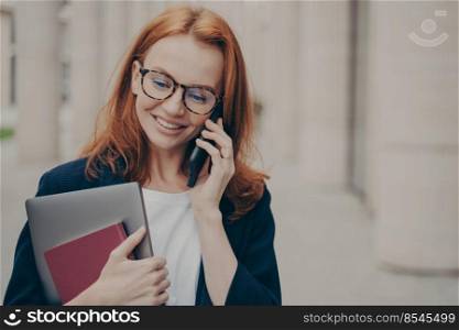 Portrait of young 30s caucasian business woman standing outdoors and talking on smartphone with client, arranging meeting with partner on mobile phone with smile on face, blurred city background. Young 30s caucasian business woman standing outdoors and talking on smartphone with client
