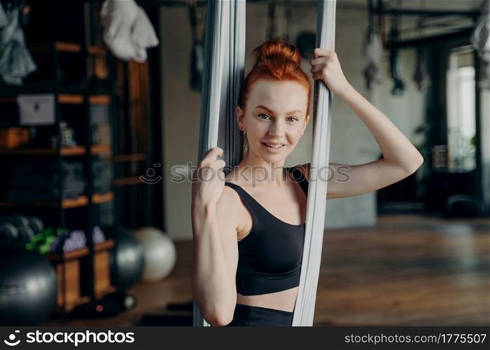 Portrait of young active and healthy female in black top sitting on fly yoga hammock and hanging over floor in gym during sports class in fitness studio background, relaxing and smiling at camera. Young active female in tracksuit sitting on fly yoga hammock