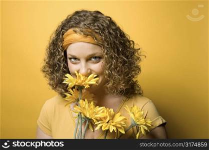 Portrait of young adult Caucasian woman on yellow background holding and smelling bouquet of flowers.