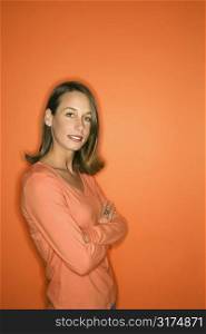 Portrait of young adult Caucasian woman standing with arms crossed making eye contact against orange background.