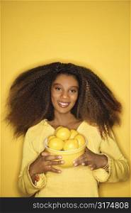 Portrait of young African-American adult woman smiling pleasantly against yellow background holding bowl of lemons.