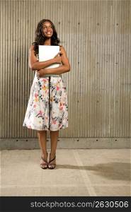 Portrait of young African American woman holding a laptop computer and standing in front of a cement wall