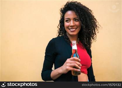 Portrait of young Afro american latin woman enjoying and drinking a bottle of beer, against yellow background. Lifestyle concept.