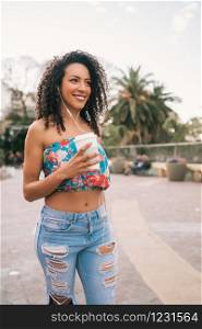 Portrait of young Afro american latin woman listening to music with earphones while holding a cup of coffee outdoors in the street.