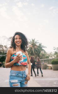 Portrait of young Afro american latin woman listening to music with earphones while holding a cup of coffee outdoors in the street.