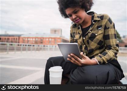 Portrait of young afro american latin woman using her digital tablet while sitting on a bench outdoors. Technology concept.
