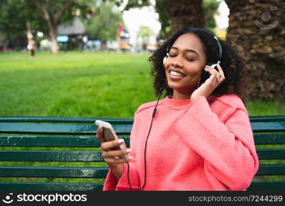 Portrait of young Afro american woman listening to music with headphones and mobile phone in the park. Outdoors.