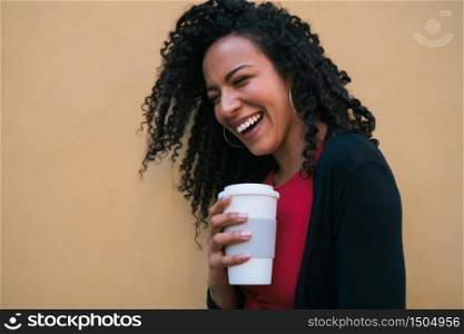 Portrait of young afro american woman looking confident and posing while holding a cup of coffee against yellow background.