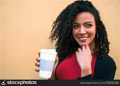 Portrait of young afro american woman looking confident and posing while holding a cup of coffee against yellow background.