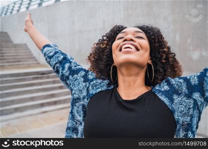Portrait of young afro american woman standing outdoors with arms raised and laughing.
