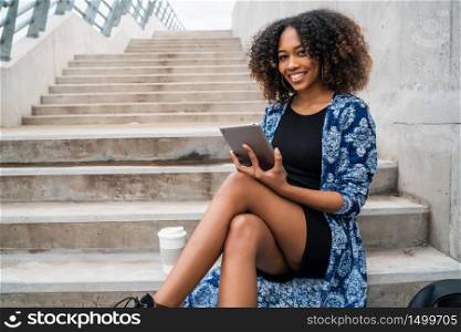 Portrait of young afro american woman using her digital tablet while sitting at stairs outdoors. Technology concept.