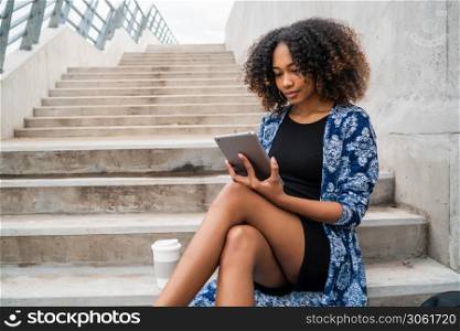 Portrait of young afro american woman using her digital tablet while sitting at stairs outdoors. Technology concept.
