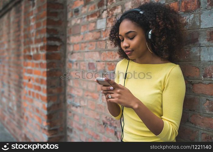 Portrait of young Afro american woman using mobile phone and listening to music with headphones in the street. Outdoors.