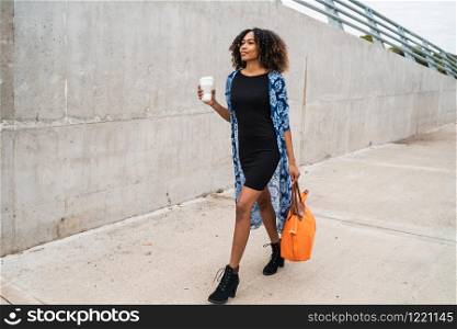 Portrait of young afro american woman walking while holding a cup of coffee and a bag outdoors.