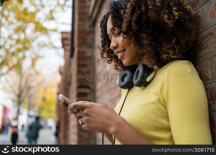 Portrait of young Afro american woman with headphones and using her mobile phone in the street. Outdoors.