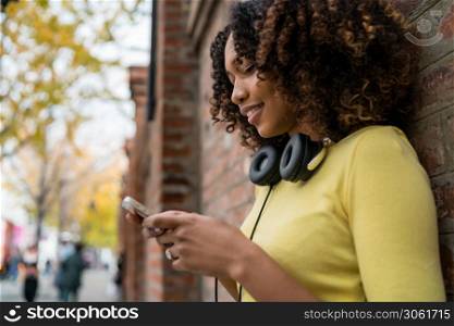Portrait of young Afro american woman with headphones and using her mobile phone in the street. Outdoors.