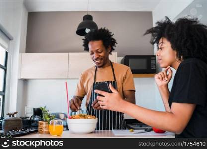 Portrait of young afro couple cooking together and using mobile phone in the kitchen at home. Relationship, cook and lifestyle concept.