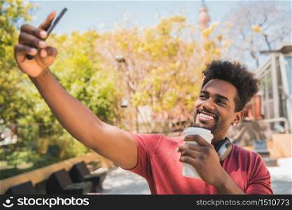 Portrait of young afro man taking selfies with his mophile phone while holding a cup of coffee outdoors. Technology concept.