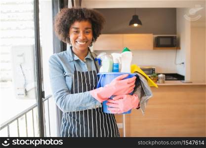Portrait of young afro woman holding a bucket with cleaning items at home. Housekeeping and cleaning concept.