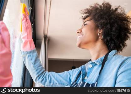 Portrait of young afro woman in gloves cleaning window with rag at home. Housework, housekeeping and cleaning concept.