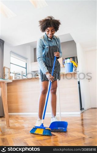 Portrait of young afro woman sweeping wooden floor with broom at home. Cleaning, housework and housekeeping concept.