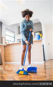 Portrait of young afro woman sweeping wooden floor with broom at home. Cleaning, housework and housekeeping concept.