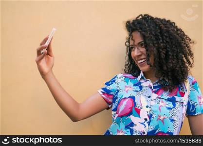 Portrait of young afro woman taking selfies with her mophile phone against yellow wall. Technology concept.