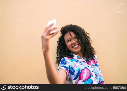 Portrait of young afro woman taking selfies with her mophile phone against yellow wall. Technology concept.