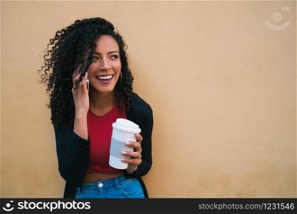 Portrait of young afro woman talking on the phone while holding a cup of coffee against yellow background. Communication concept.