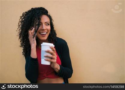 Portrait of young afro woman talking on the phone while holding a cup of coffee against yellow background. Communication concept.
