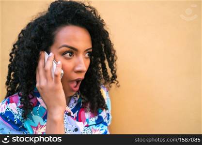 Portrait of young afro woman talking on the phone with shocked expression against yellow background. Communication concept.