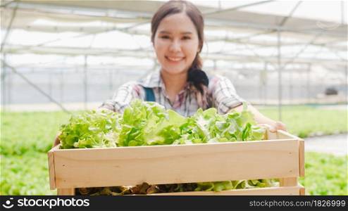Portrait of young Asia lady farmer holding basket of green oak looking at camera and smile in hydroponics vegetable farm in morning. Agriculture organic for health, Vegan food, Small business concept.