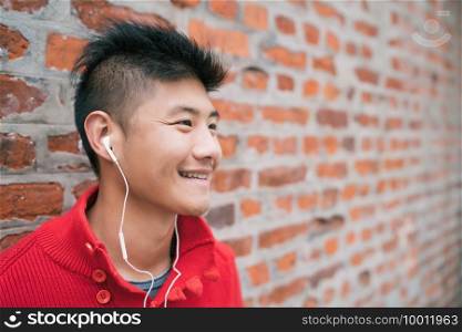 Portrait of young Asian boy listening to music with earphones outdoors against brick wall. Urban concept.