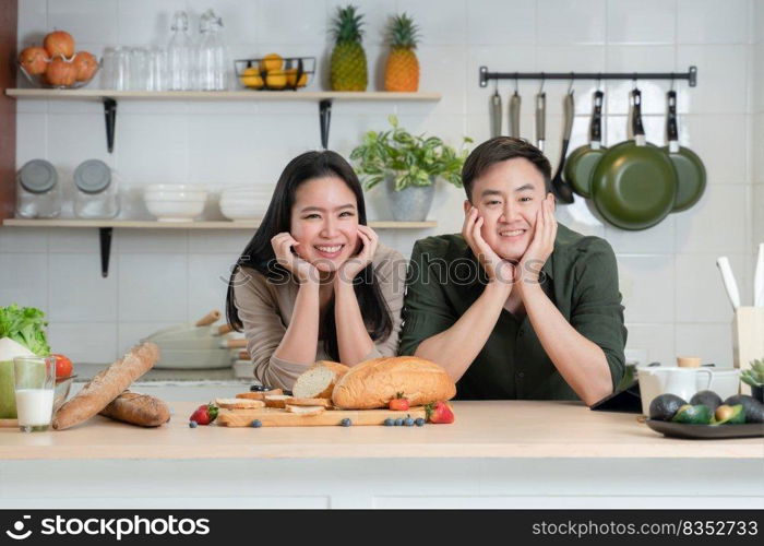 Portrait of young Asian couple smiling, placing hands on chin, looking at camera in the kitchen, happy beautiful wife and handsome husband cooking and prepare healthy breakfast food together at home