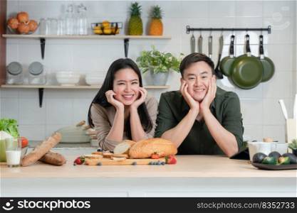 Portrait of young Asian couple smiling, placing hands on chin, looking at camera in the kitchen, happy beautiful wife and handsome husband cooking and prepare healthy breakfast food together at home
