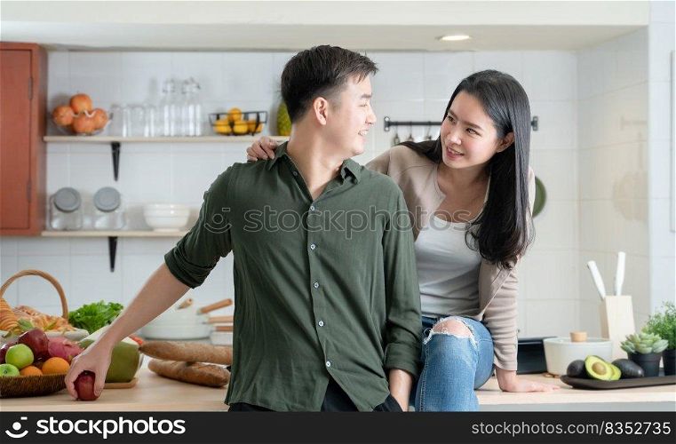 Portrait of young Asian couple standing in the kitchen, happy romantic lover smiling and looking at each other, beautiful woman hugging handsome man from behind while cooking together at home